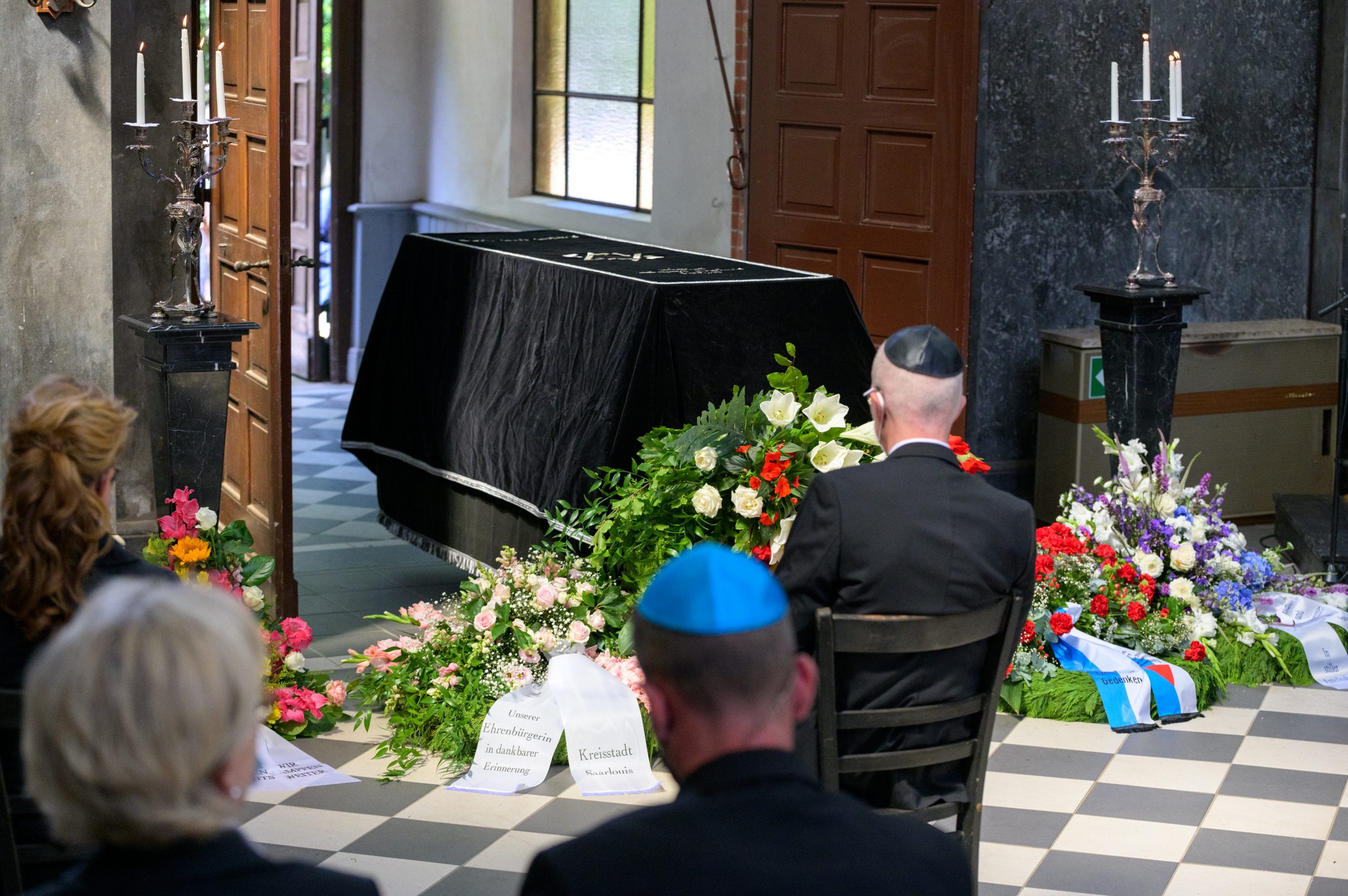 Der mit einem Tuch bedeckte Sarg von Esther Bejarano steht vor Blumenkränzen und Trauergästen in der Kapelle.